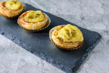 Three homemade mini tarts with golden crust and creamy topping placed neatly on a black slate board with a clean textured background. Perfect for bakery promotions, dessert menus