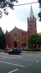 Obraz premium A brick cathedral with a tall spire, framed by trees and viewed from a busy city street on a cloudy day