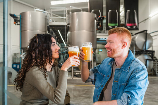 Joyful couple enjoying a beer tasting experience, clinking glasses inside a modern microbrewery production plant