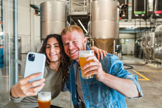 Happy couple visiting a modern brewery, drinking craft beer, and taking a selfie on their smartphone for social media