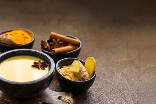 Traditional Ayurvedic golden milk latte in a dark mug, surrounded by its wholesome ingredients&mdash;fresh turmeric root, ground cinnamon, ginger slices, and honey