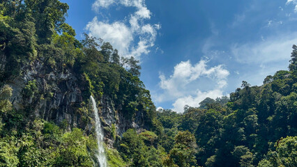 Thin waterfall descends cliff against bright sky
