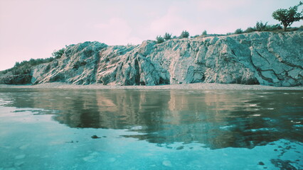 Clear waters reflect rugged cliffs along the beach, while lush greenery tops the rocky landscape. The scene is tranquil, capturing a peaceful moment at sunrise with soft colors.