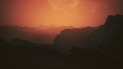 A vibrant sunset casts hues of orange and red over jagged mountain peaks. The landscape is tranquil, evoking a sense of peace as day transitions into night. Shadows deepen in the valleys.