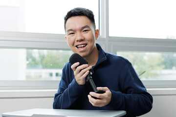 Smiling young man holding a microphone while recording a podcast in a classroom. Represents student media, communication, and creative expression in education.