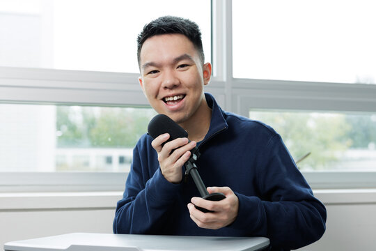 Smiling young man holding a microphone while recording a podcast in a classroom. Represents student media, communication, and creative expression in education. - Powered by Adobe