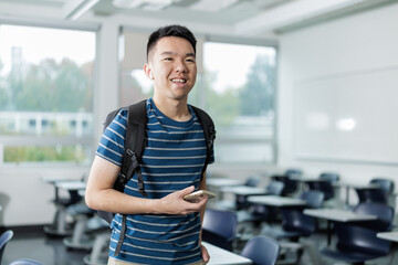 Young man with backpack holding a smartphone and earphones in a modern classroom, symbolizing digital learning and connected student life.