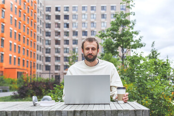 bearded man in light sweatshirt works on laptop outdoors at wooden table with coffee headphones...