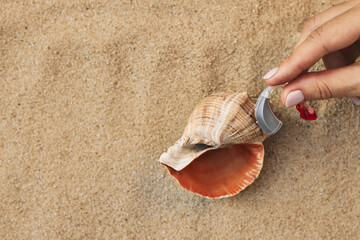 Hearing aid sound clarity, concept. Exploring the synergy between nature and technology, a hand holds a hearing aid near seashells on soft sand