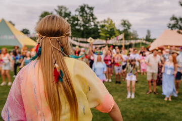 Naklejka premium A woman with long hair and feathers in her hair stands facing a cheerful outdoor crowd at a colorful summer festival or event, seen from behind.