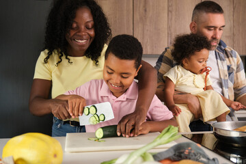 Mother teaching son to cut cucumber while family cooks in kitchen