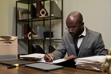 Black man sitting at desk working with documents and writing on paper, surrounded by stacks of files and folders, wearing glasses and formal suit in office setting