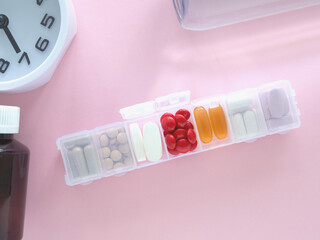 Top view, the medicine box is open, various white and dark brown pills or tablets in different compartments and clock on pink table. Concept daily routines and medication management supplements.