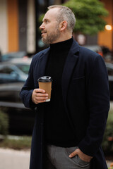 Smiling businessman in coat holding coffee to go on city street
