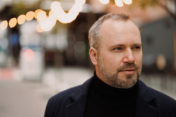 Bearded man in coat and turtleneck standing on blurred city street