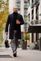 Adult businessman in coat holding takeaway coffee while walking on street