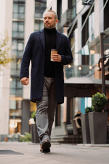 Confident man in coat holding coffee to go while walking on street