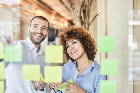 Two happy colleagues brainstorming with post-its on glass pane