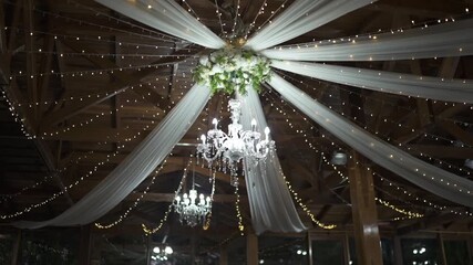A beautiful low-angle shot of a wedding venue's ceiling, decorated with a crystal chandelier, white fabric drapery, and twinkling fairy lights for a magical, romantic ambiance. - Powered by Adobe