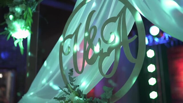 Artistic close-up of a wooden circular sign displaying the initials V and A, symbolizing the bride and groom, surrounded by flowers and white drapery under romantic green lighting at wedding reception