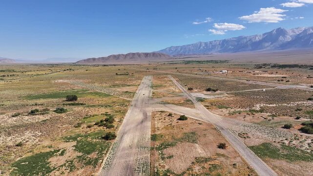 Drone pulls back and ascends over the former Manzanar airfield in Inyo County California revealing faint concrete runway lines desert scrub and the Eastern Sierra backdrop on a clear summer day.