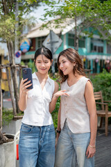 Pretty asian woman holding phone taking picture with friend and standing at sidewalk by street road.