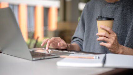 Close up of a man's hands holding coffee cup typing on laptop aside notebook while sitting at table.