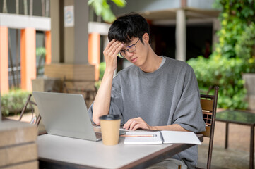 Glasses asian man holding head suffering with study aside laptop and notebook while sitting at table