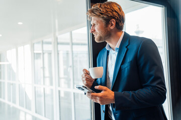 Manager in suit holding coffee and smartphone at office window