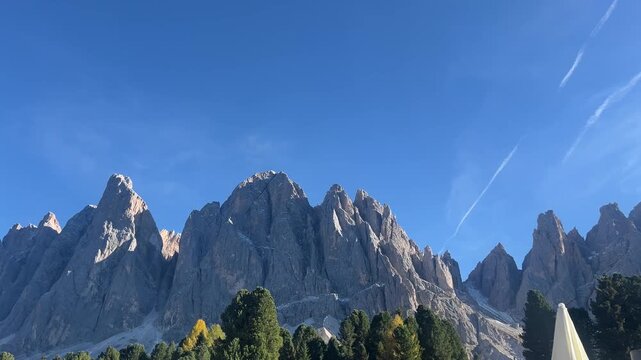 Close-up view of the Odle Peaks in Alpe di Funes, Dolomites, Italy. Dramatic rocky cliffs rise above pine forests under a bright blue sky - pure alpine majesty.