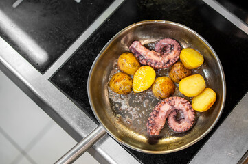 Pan-fried octopus tentacles with golden potatoes in a stainless steel skillet on stovetop. Mediterranean seafood dish, rustic home cooking, close-up food photography.