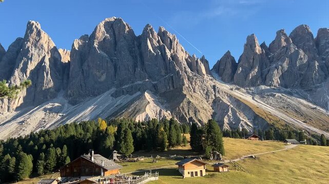 Majestic view of the Odle Peaks in Alpe di Funes, Dolomites, Italy. Wooden huts, green meadows, and dramatic rocky mountains glowing in the warm evening light.