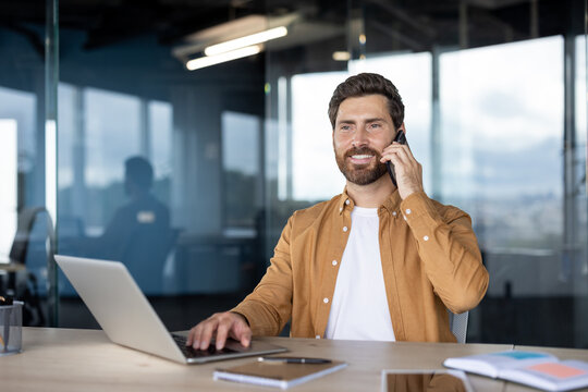 Businessman on a phone call smiling while typing on a laptop at a modern glass-partitioned desk, multitasking in a bright office, confident and productive during the workday