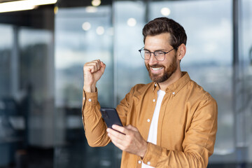 Enthusiastic man smiling widely and raising his fist in an office environment, celebrating good...