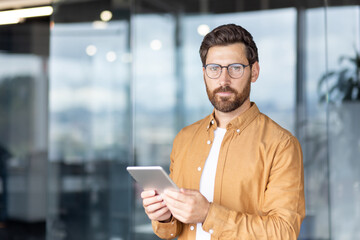 Confident bearded man with glasses standing in a modern office, holding a tablet and engaging with...