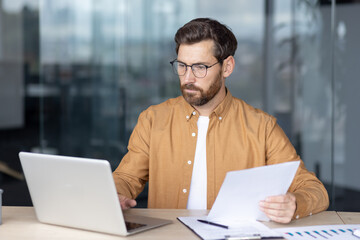 Bearded businessman with glasses intensely working on a laptop, holding documents and analyzing...