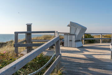 A single beach chair stands on a wooden platform overlooking the calm sea. The warm light and gentle breeze evoke pure relaxation.