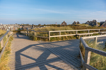 A wooden walkway stretches through the dunes with views of coastal houses and a lighthouse. Shadows fall across the boards in the soft evening light.