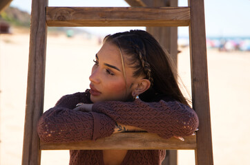 A beautiful young woman in a brown dress rests her arms on the steps of the wooden ladder of the lifeguard hut on the beach. The woman is sad and depressed and is going through a difficult time.