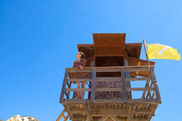 A beautiful young woman in a brown dress looks out to sea from the wooden lifeguard tower with a yellow flag. In the background is a blue sky.