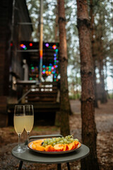 Two flutes of champagne and a plate of sliced peaches and grapes on a small table outside a rustic cabin. Colorful string lights decorate the stairs in the blurred background, hinting at a celebration