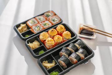 Assorted sushi rolls in black takeout boxes with soy sauce, ginger, and chopsticks on a white table, featuring salmon, baked, and seaweed maki, representing Japanese cuisine and modern food delivery