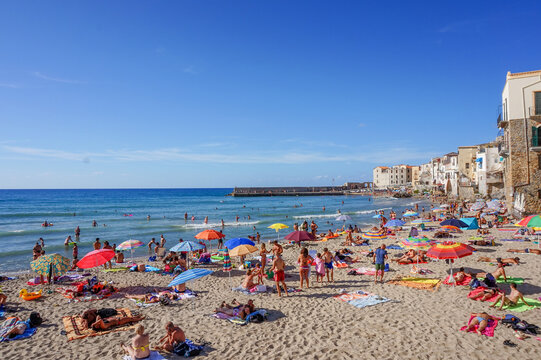 A busy summer day on the sandy beach of Cefalù, Sicily, with tourists sunbathing next to the historic old town buildings - Powered by Adobe