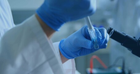 Lab tech wearing lab coat pipetting blue solution into test tube with micropipette at lab bench