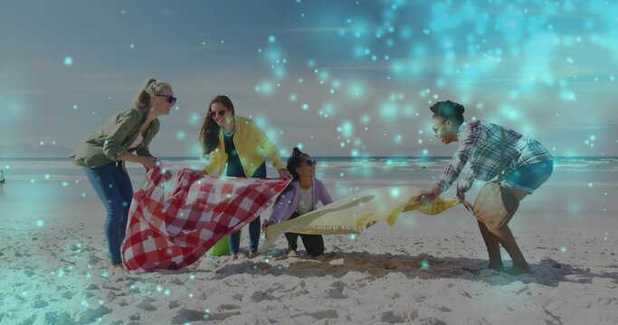 Four friends spreading checkered blanket and gold blanket on sandy beach, with turquoise glow