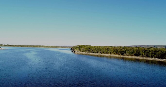 Fototapeta Showing water surface rippling under blue sky at lake, with curving wooded shoreline, faint birds