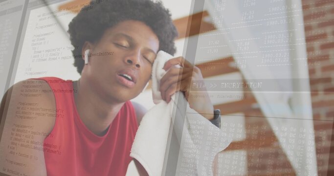 Leaning man wearing red sleeveless top pressing white towel to temple in home gym, code overlay