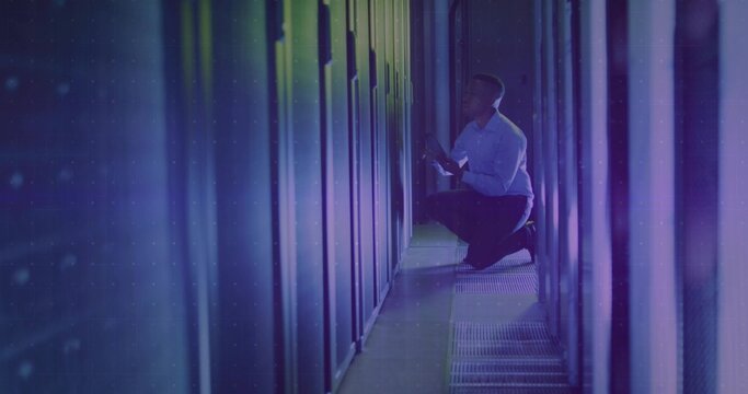 Kneeling African American technician inspecting server rack with LEDs in data center, with tablet