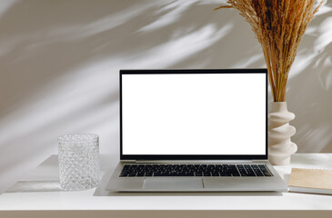 Laptop mockup with white screen and vase with pampas grass and glass of water, notepad and aesthetic shadows. Minimal desktop, laptop for web site promotion, autumn design, home office or branding