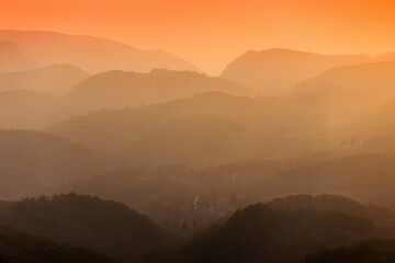 Layered mountains and hills, fading into an atmospheric and peaceful orange sunset haze over a valley village